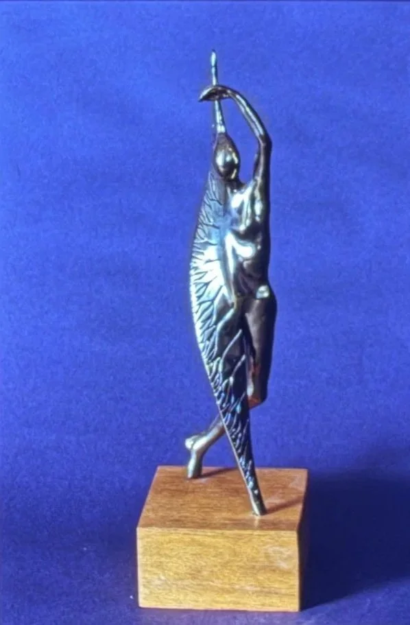 Bronze dancer enclosed within a vertical leaf-like form, reaching upward with one arm while decorative ridges flow around the body like protective wings.