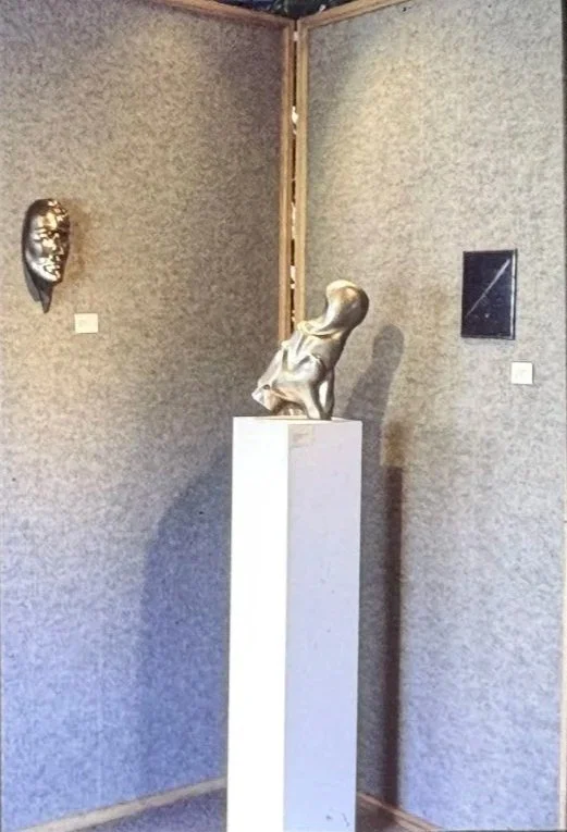 Gallery installation showing a bronze figurative sculpture displayed on a tall pedestal within a gallery space, surrounded by wall-mounted works and exhibition lighting.