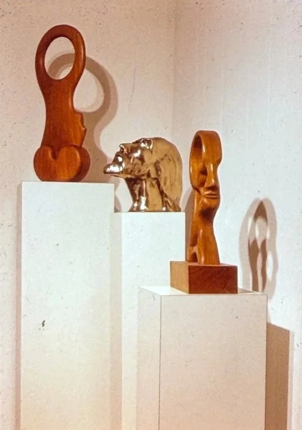 Several polished wood sculptures displayed on white pedestals inside an exhibition space.