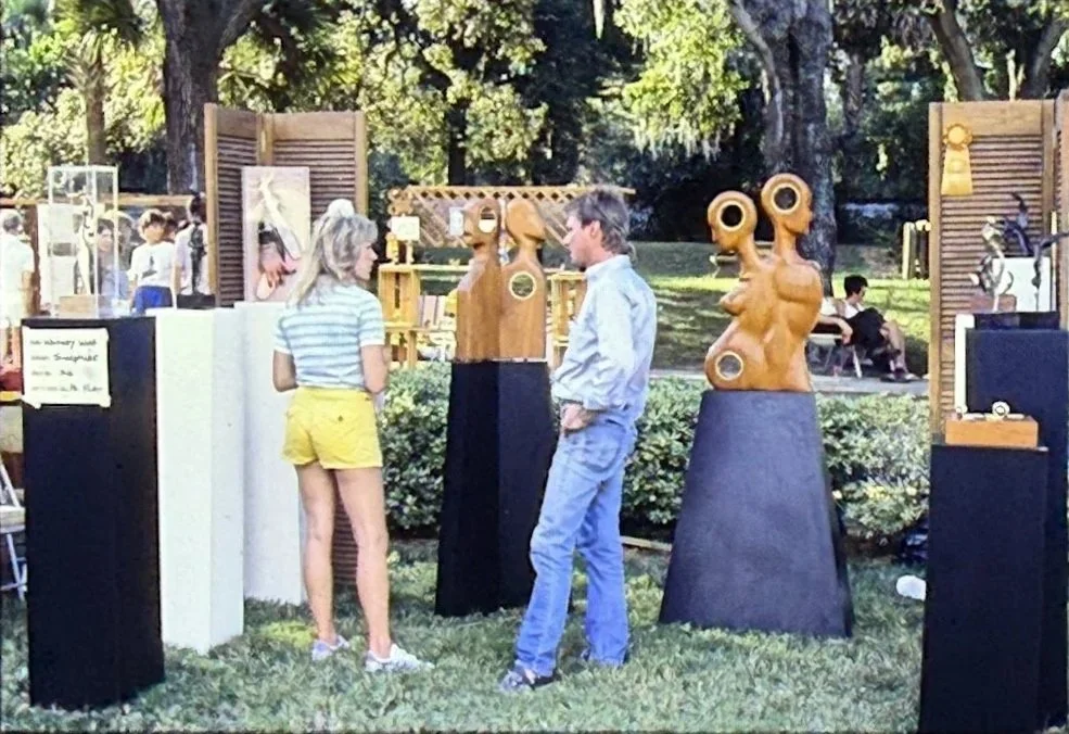 Visitors viewing wooden sculptures arranged throughout an outdoor art fair installation.