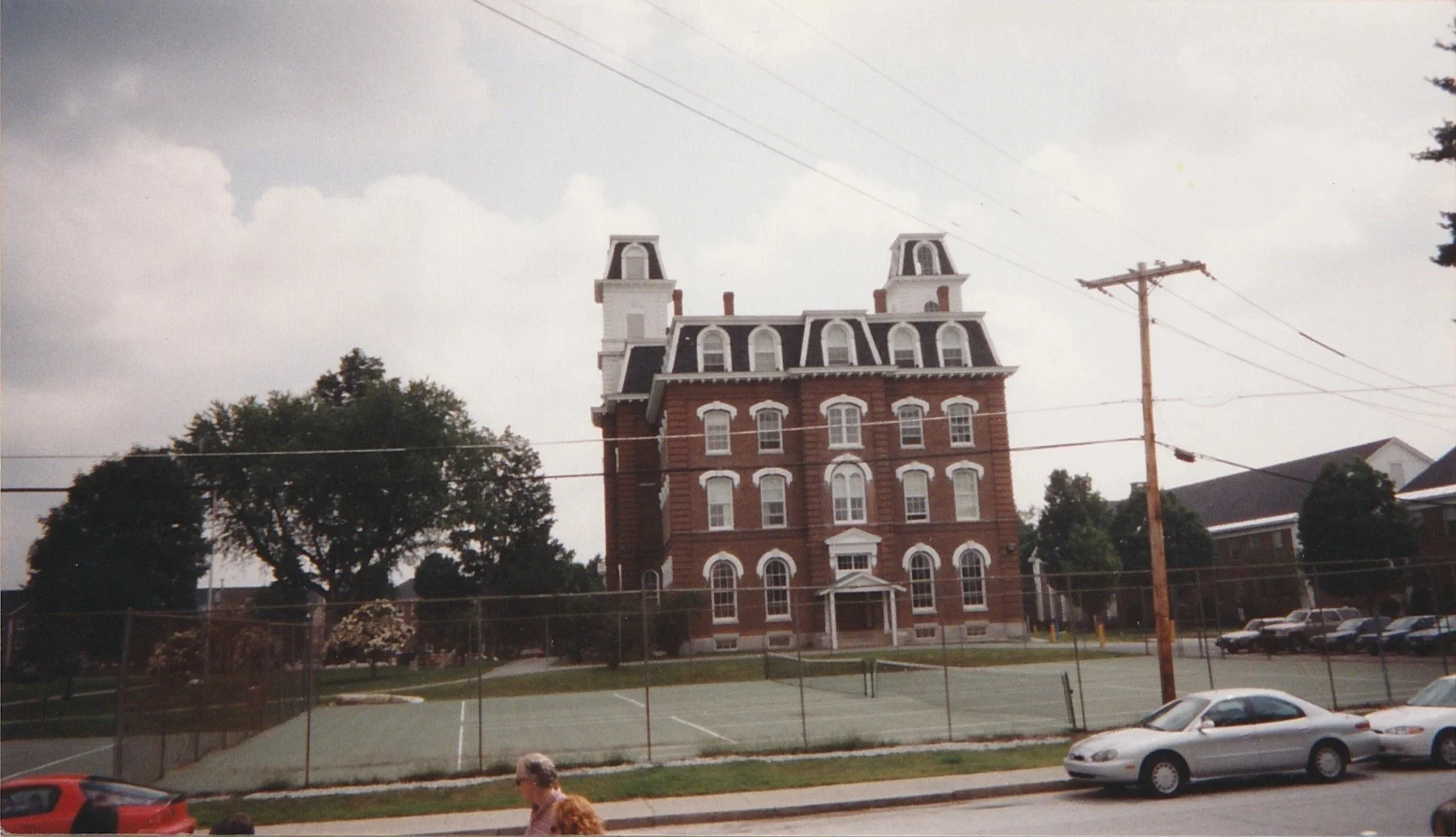 Large red-brick institutional building with twin towers and white trim viewed from across a street under cloudy skies.