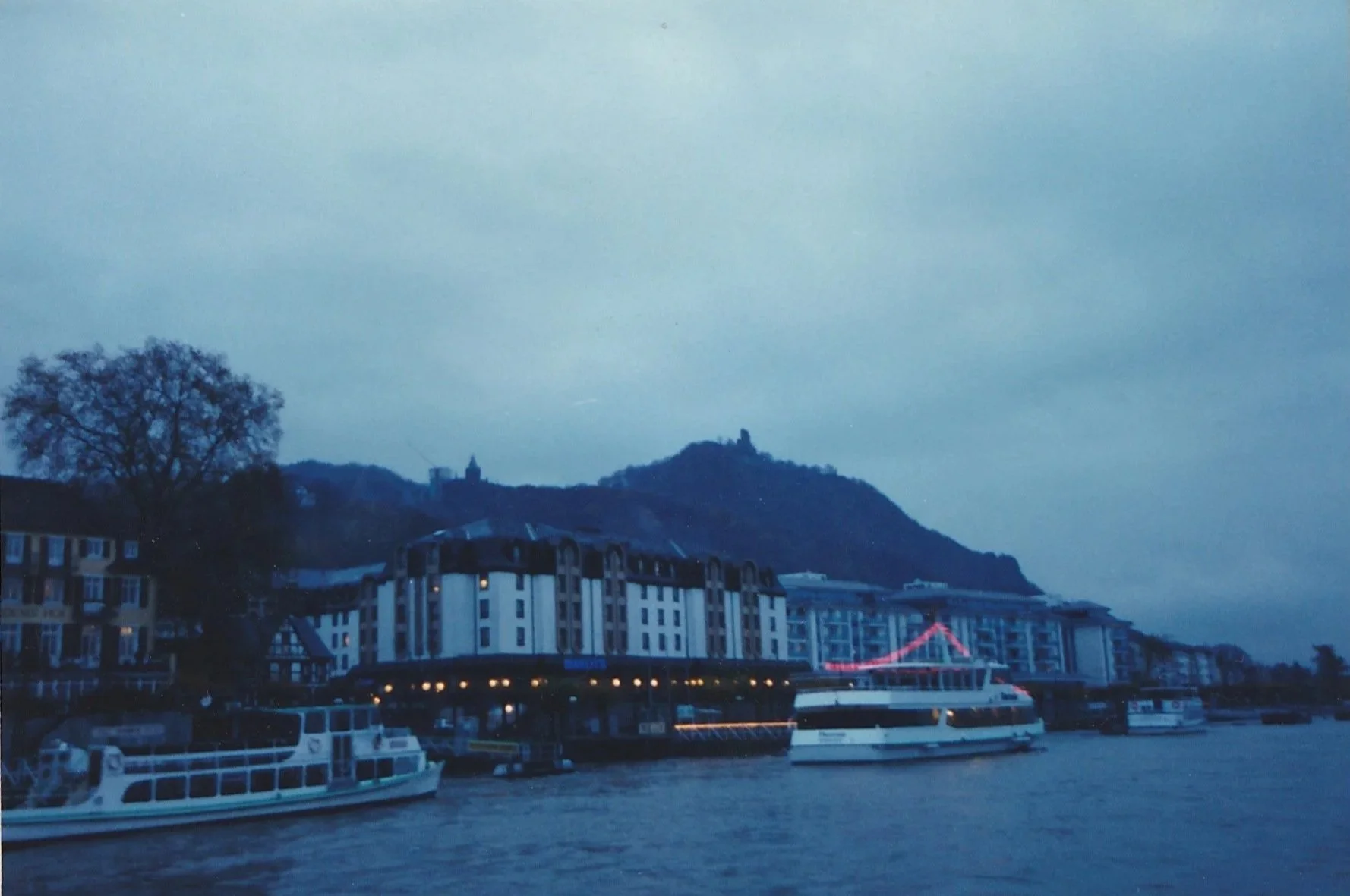 Waterfront cityscape with boats moored along a river beneath overcast skies.