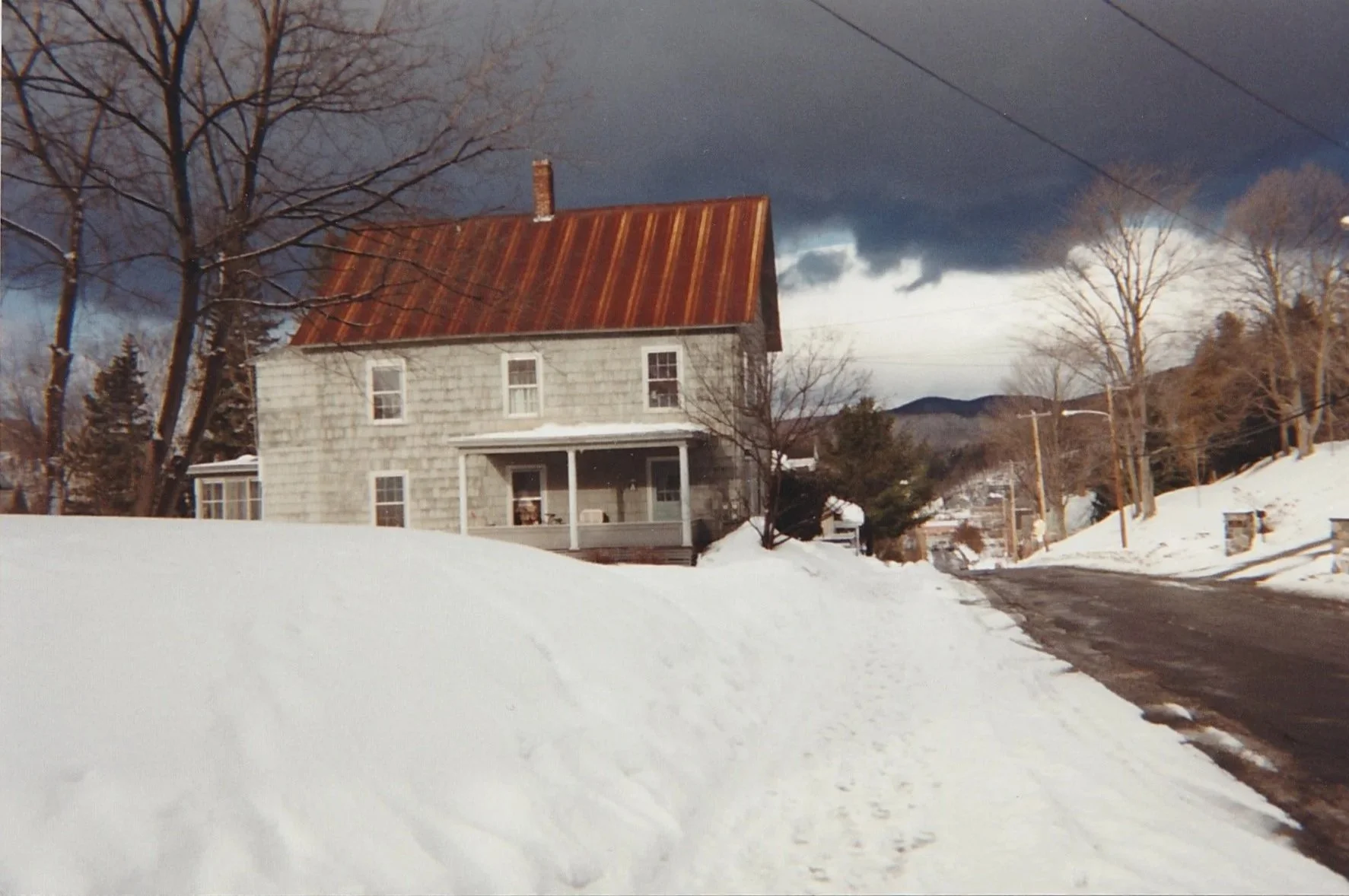 Grey brick farmhouse with a rusted tin roof beside a snowbank under dramatic storm clouds.