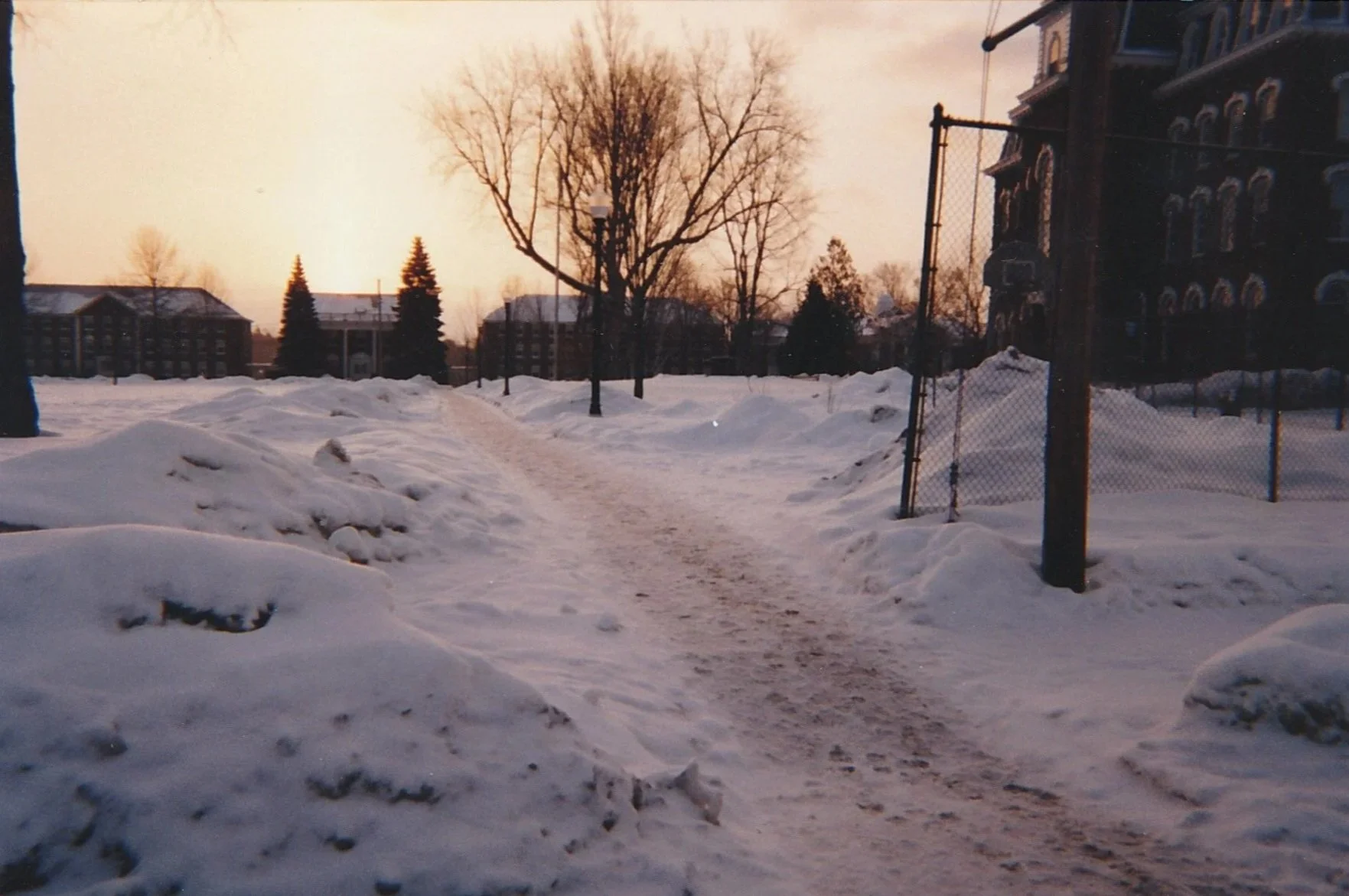 Narrow path carved through deep snowdrifts leading toward distant buildings beneath a pastel evening sky.