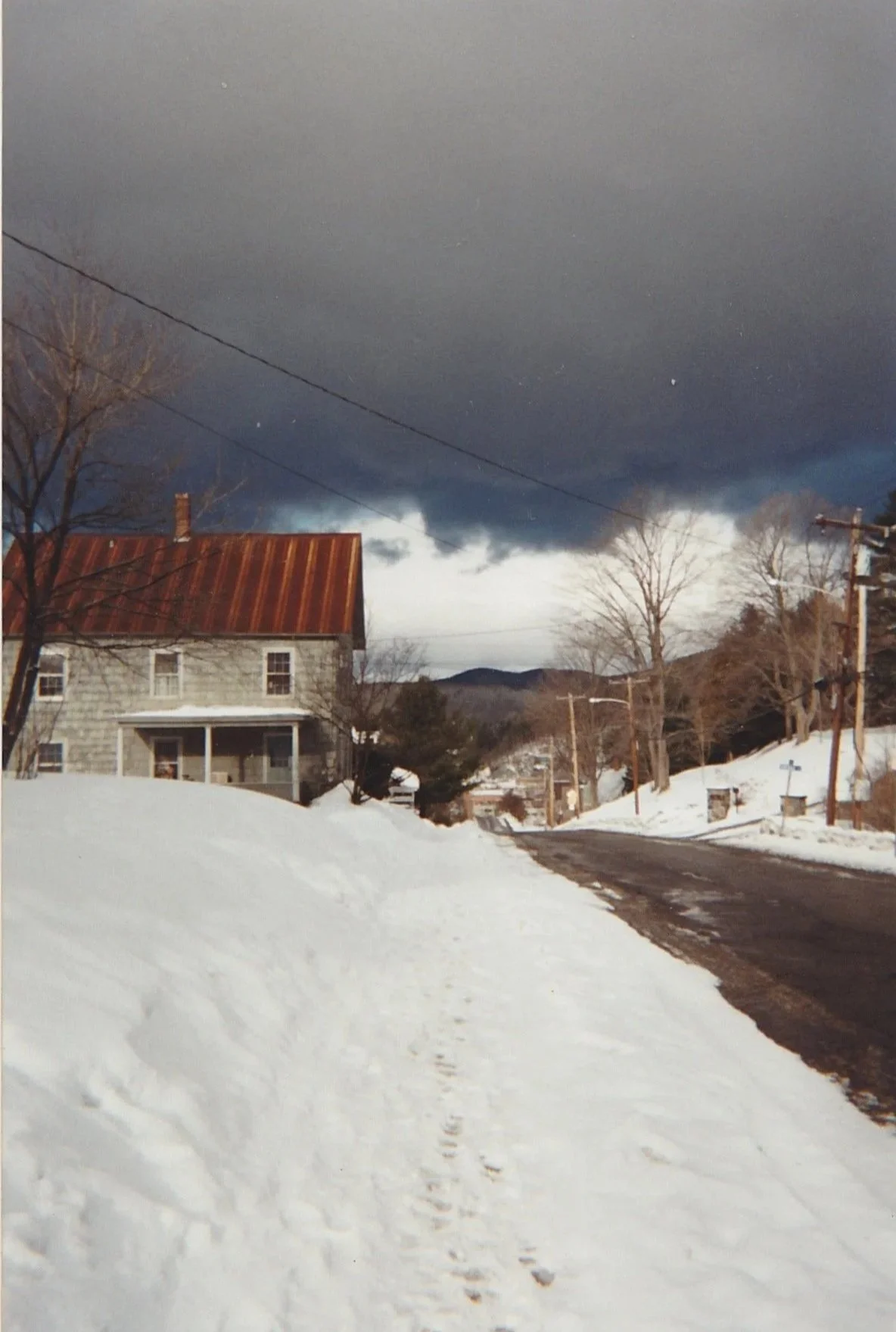 Country road bordered by snowbanks leading toward a farmhouse beneath dark storm clouds.