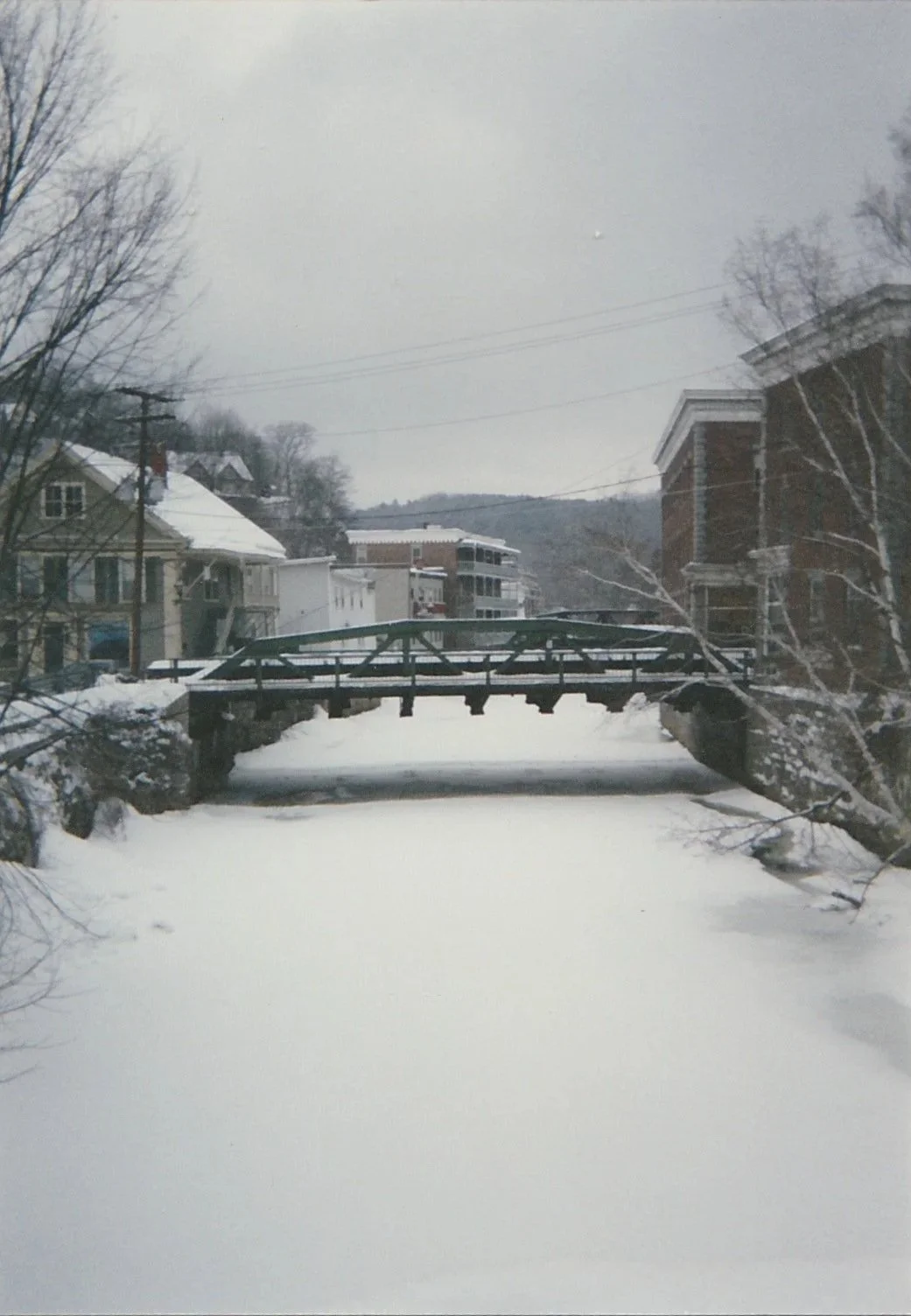 Snow-covered canal crossed by a small bridge, framed by winter trees and historic buildings.