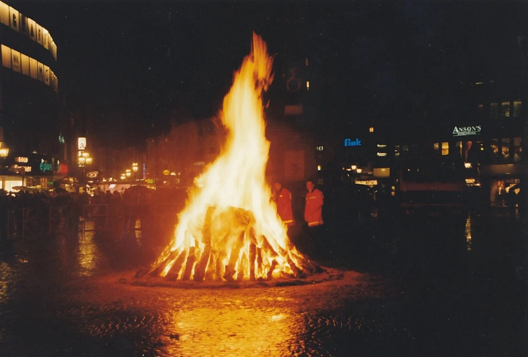 Large bonfire burning at night in a public square, its flames reflected on wet pavement.