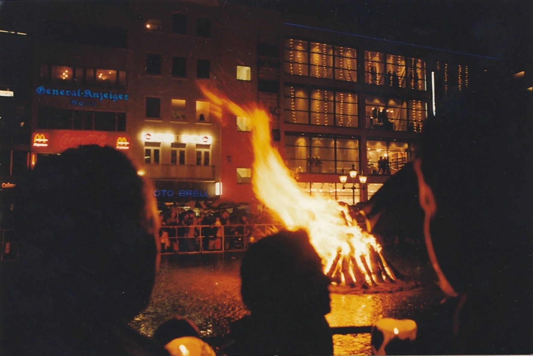 Crowd gathered around a nighttime bonfire in a city square, watching flames rise against illuminated buildings.