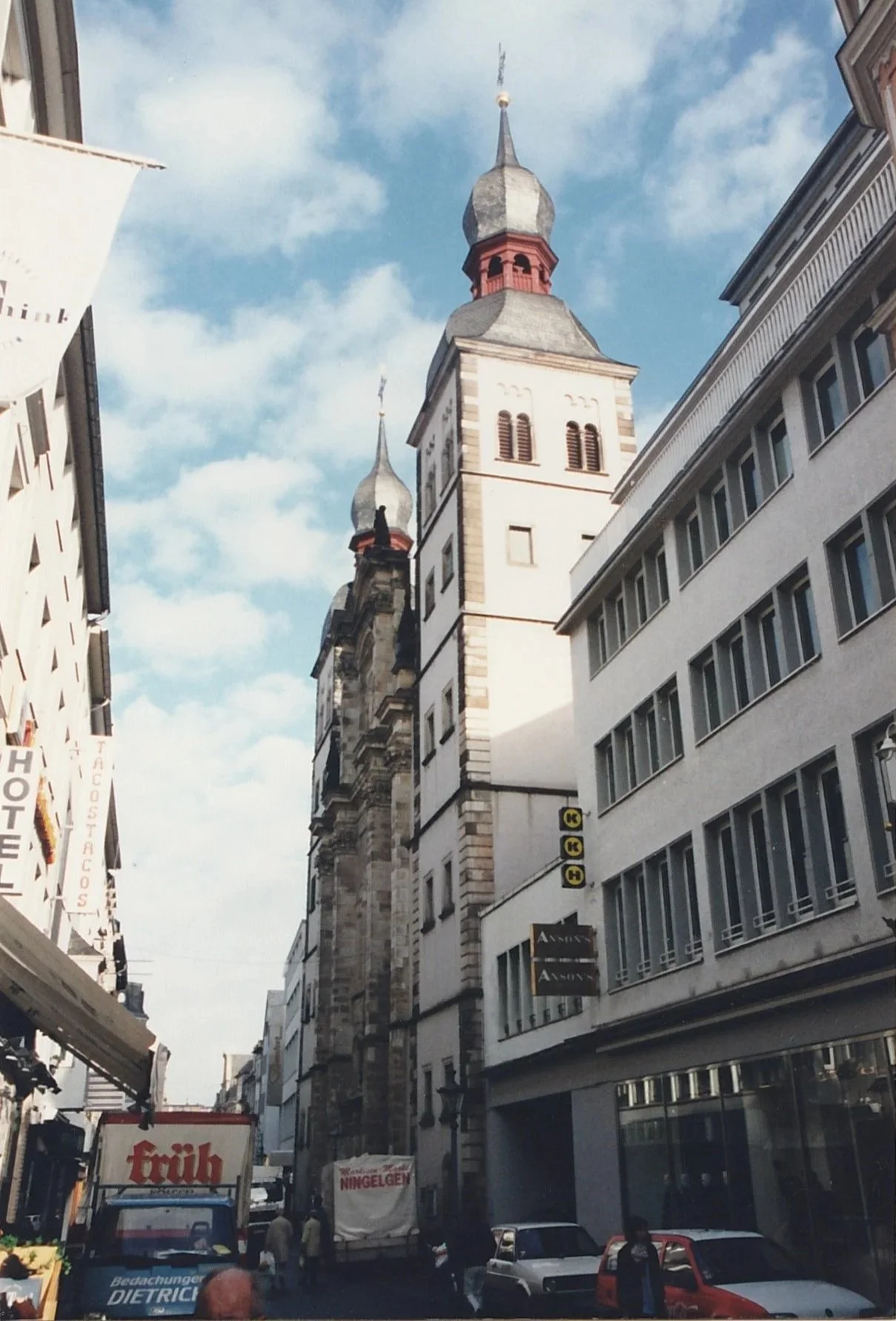 Historic European street framed by modern buildings, leading the eye toward a tall church tower crowned with distinctive onion-shaped domes.