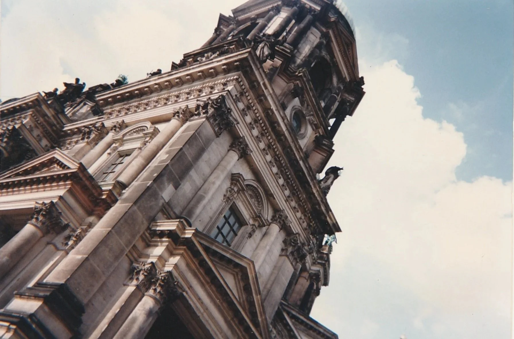 Dramatic low-angle photograph of a monumental Baroque cathedral, its richly ornamented stone columns, cornices, and sculptural details rising toward the sky. The towering architecture fills the frame