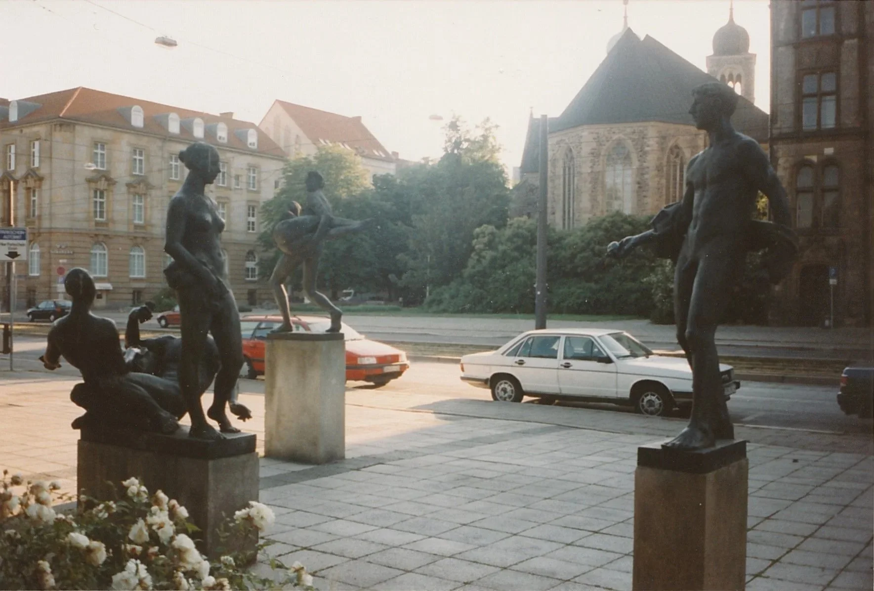 Bronze figurative sculptures stand on stone pedestals in an urban plaza, their dark silhouettes illuminated by soft afternoon light.