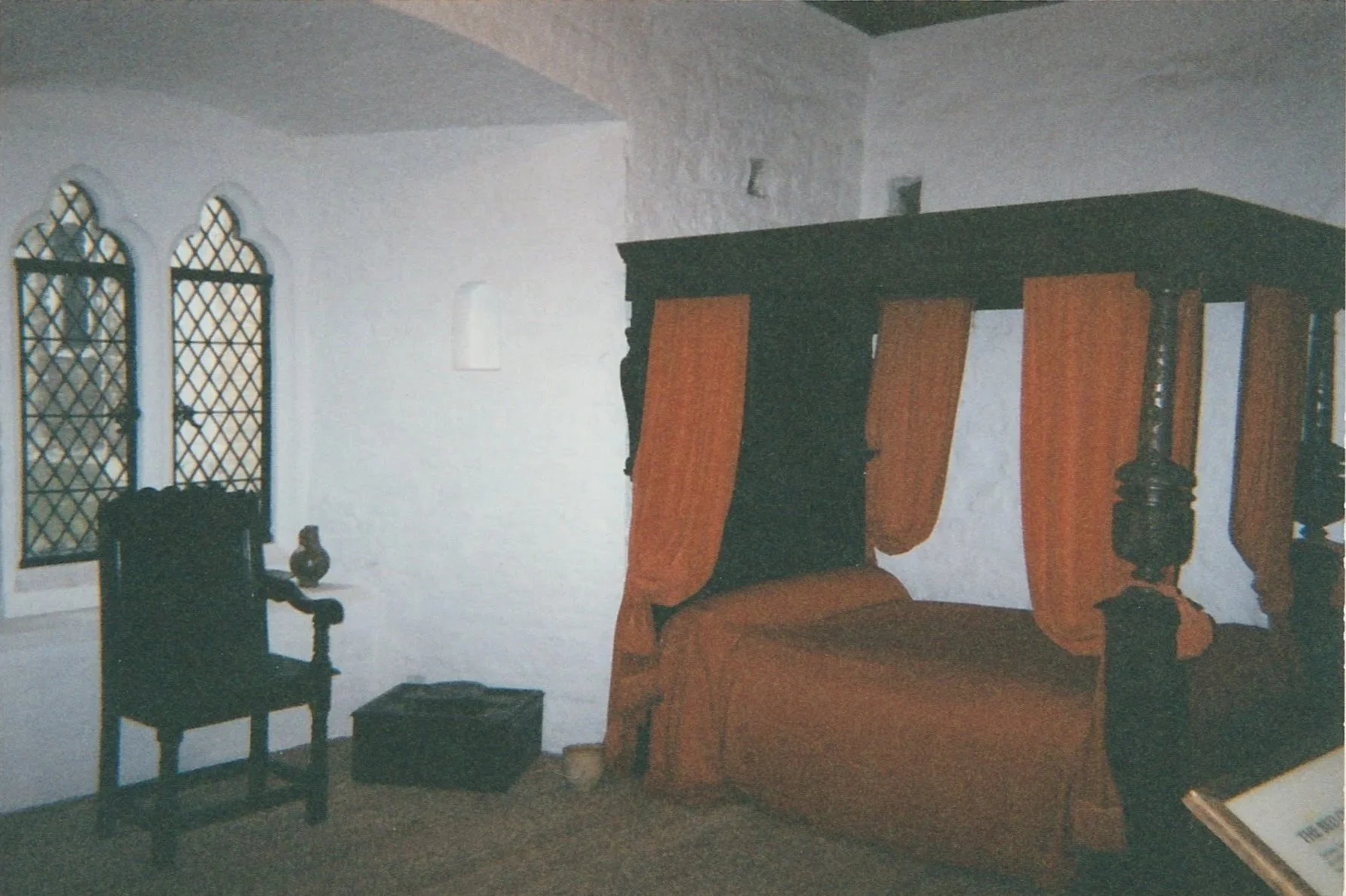 A simple historic room contains a dark wooden canopy bed draped with warm red curtains beside leaded-glass windows.
