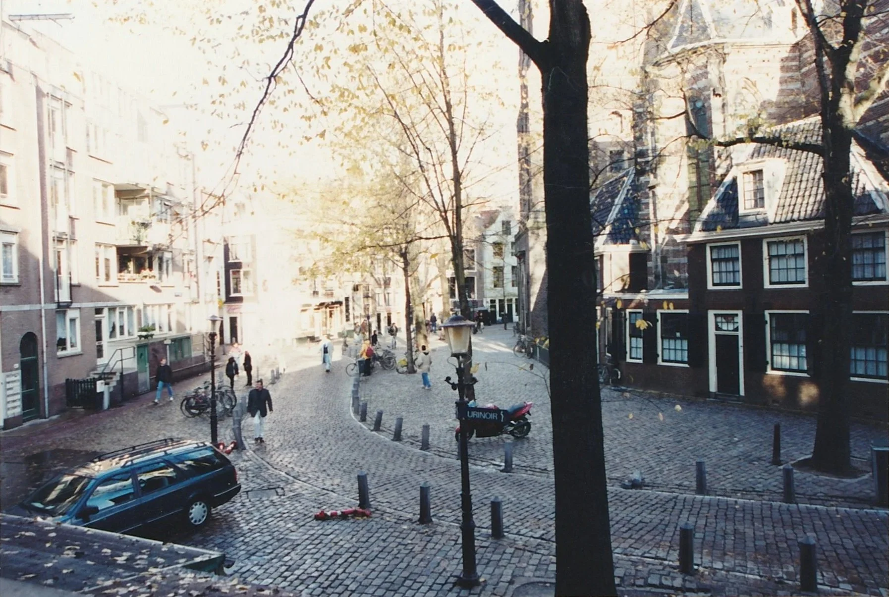 Sunlight filters through bare trees onto a cobblestone square surrounded by narrow canal houses and historic buildings. Pedestrians, bicycles, and parked cars animate the peaceful neighborhood scene.