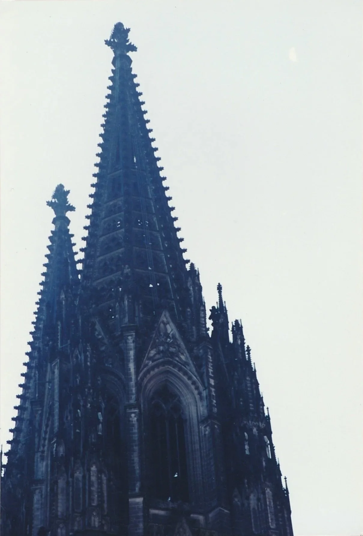 A towering Gothic church spire rises sharply into a pale sky, its dark stone surface covered with intricate tracery and decorative ornament. 