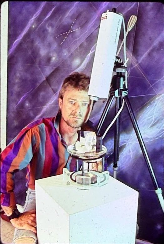 Whitney Wolf II is wearing a colorful shirt, and sitting in front of one of his purple paintings. In front of him are his science themed sculptures. EST 1980s