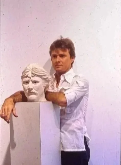 Whitney Wolf II leaning against a pedestal that holds a self-portrait sculpture head of himself. EST 1970s