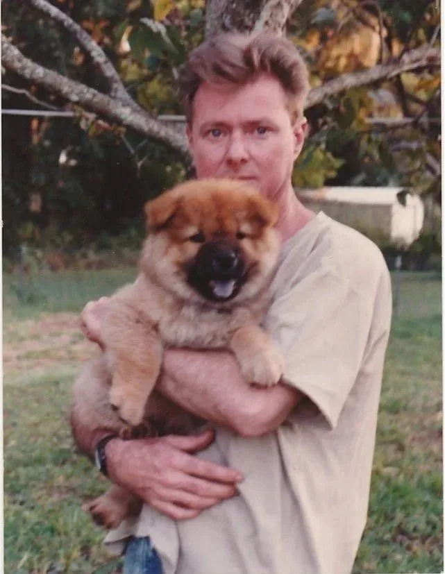 Whitney Wolf II outside in the grass holding a dog. There is a tree behind him and maybe a shed. EST 1970s