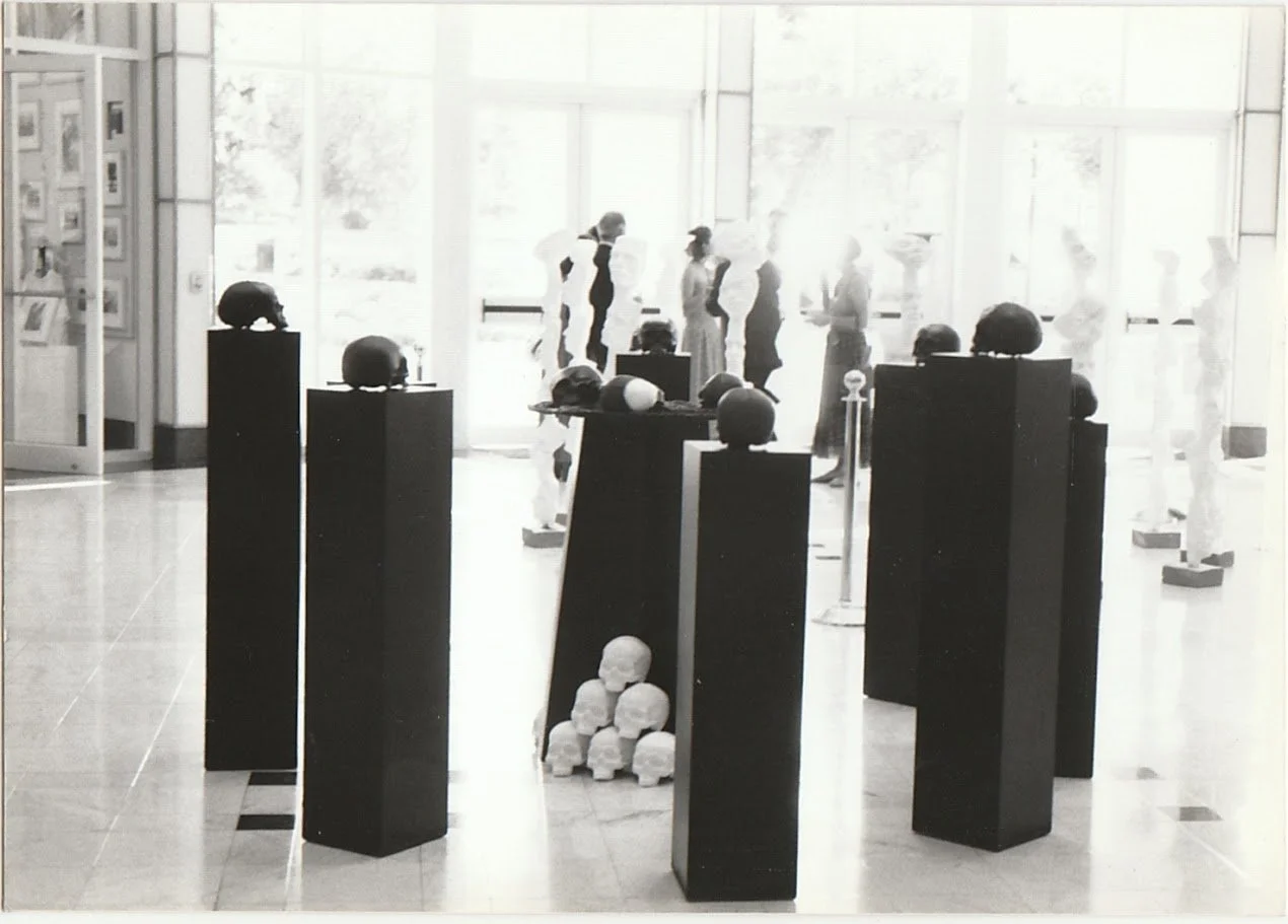 Multiple skull sculptures are positioned throughout a gallery space, creating a layered arrangement of forms at varying heights and distances. A pyramid of white skulls sits at the base of the pedestals. 