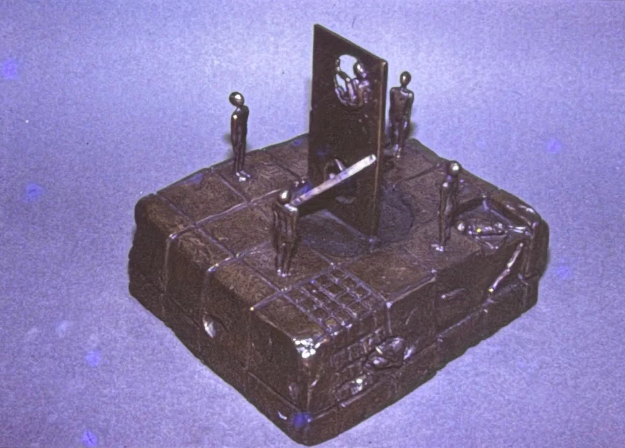 Architectural bronze sculpture resembling a miniature stage or monument. Small human figures stand around a central panel and staircase-like structure on a textured base.