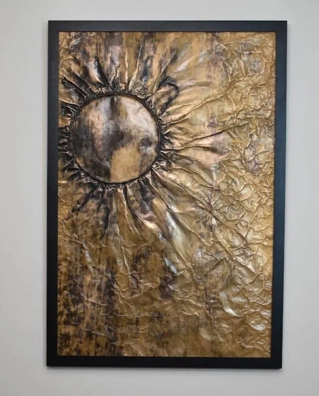 A dark orb radiates through a textured field of gold and bronze. Displayed framed on a white wall.
