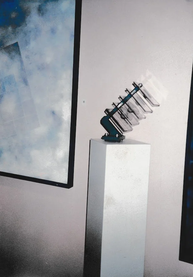 Small abstract sculpture displayed on a pedestal, featuring stacked transparent elements and metal supports against a minimalist gallery wall.