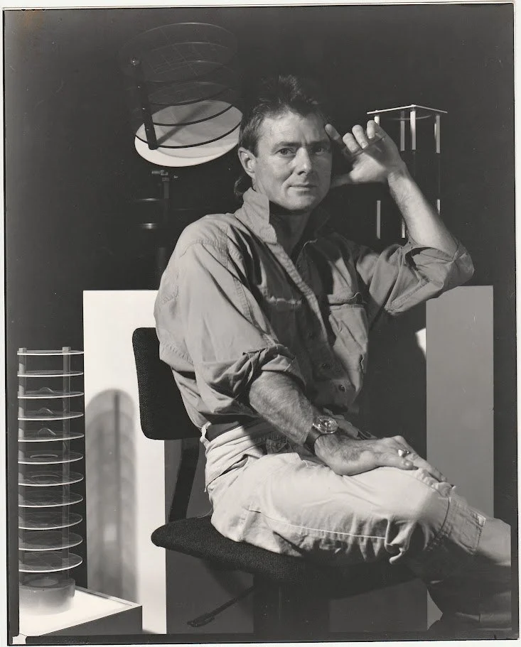 Whitney Wolf II in a black-and-white photo where he is sitting. He is resting his left hand on his temple, and his right hand on his knee. Wolf is surrounded by the scientific sculptures that he created. EST 1980s.