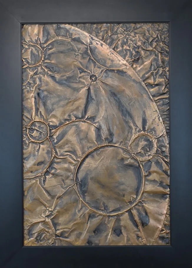 Circular forms drift across a fractured metallic field. Shown close up in a black frame.
