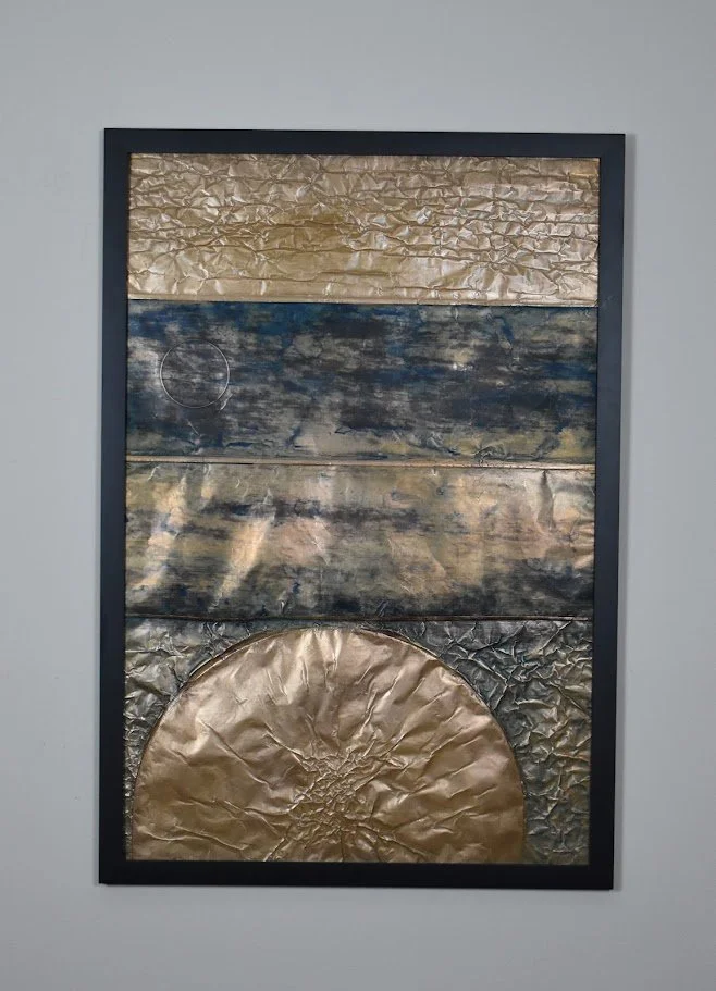 A golden orb rises beneath layered bands of dark blue and metallic gold. Displayed framed on a white wall.