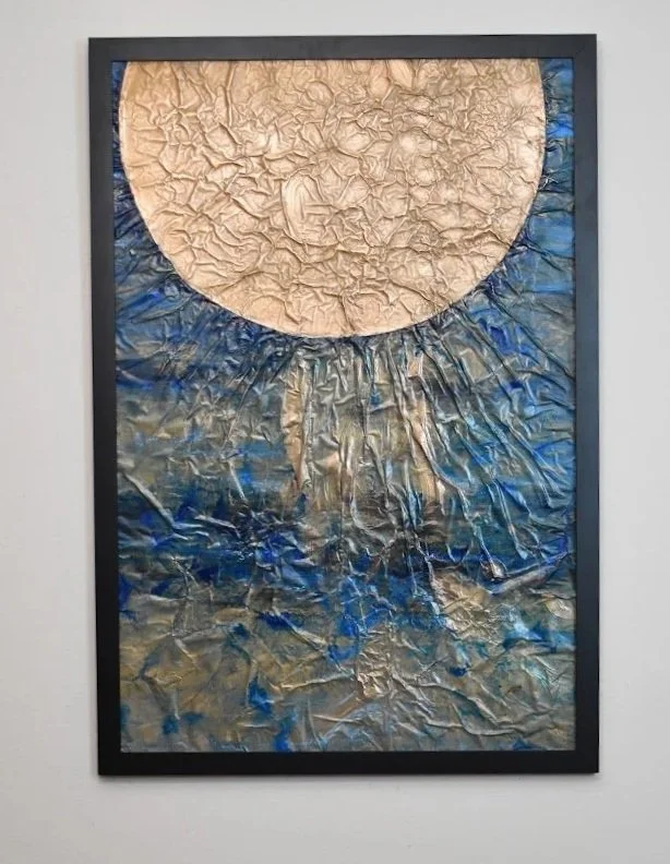 A pale orb radiates over turbulent blue currents. Displayed framed on a white wall.