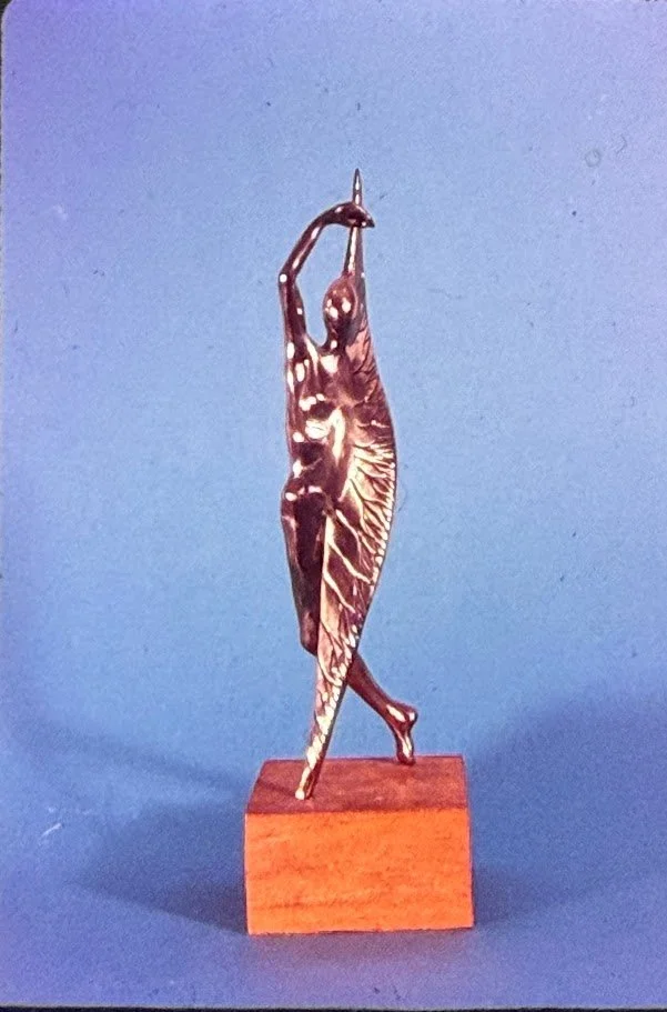 Delicate bronze figure enclosed within a leaf-like vertical form, arms lifted overhead as if emerging from a protective shell.