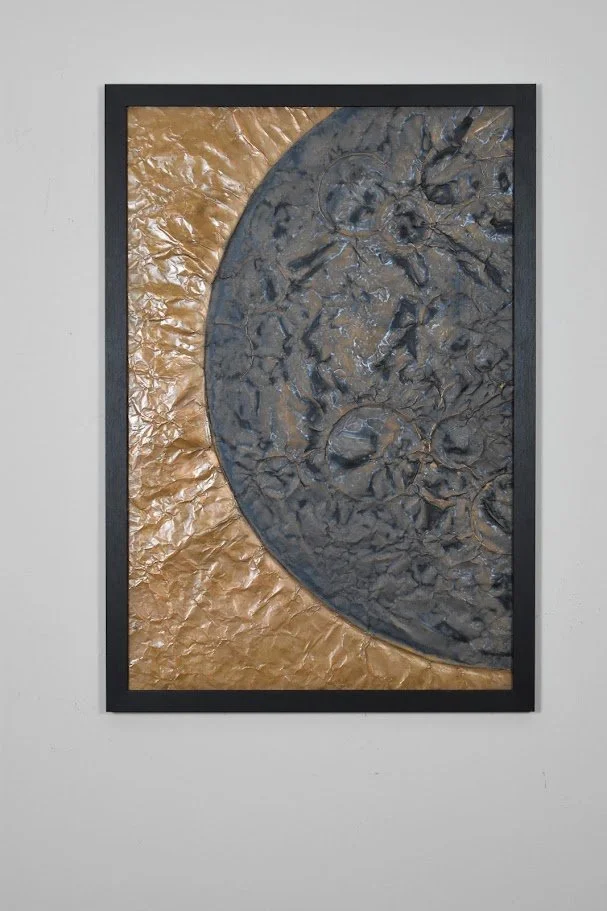 A cratered dark world curves across a luminous golden field. Displayed framed on a white wall.