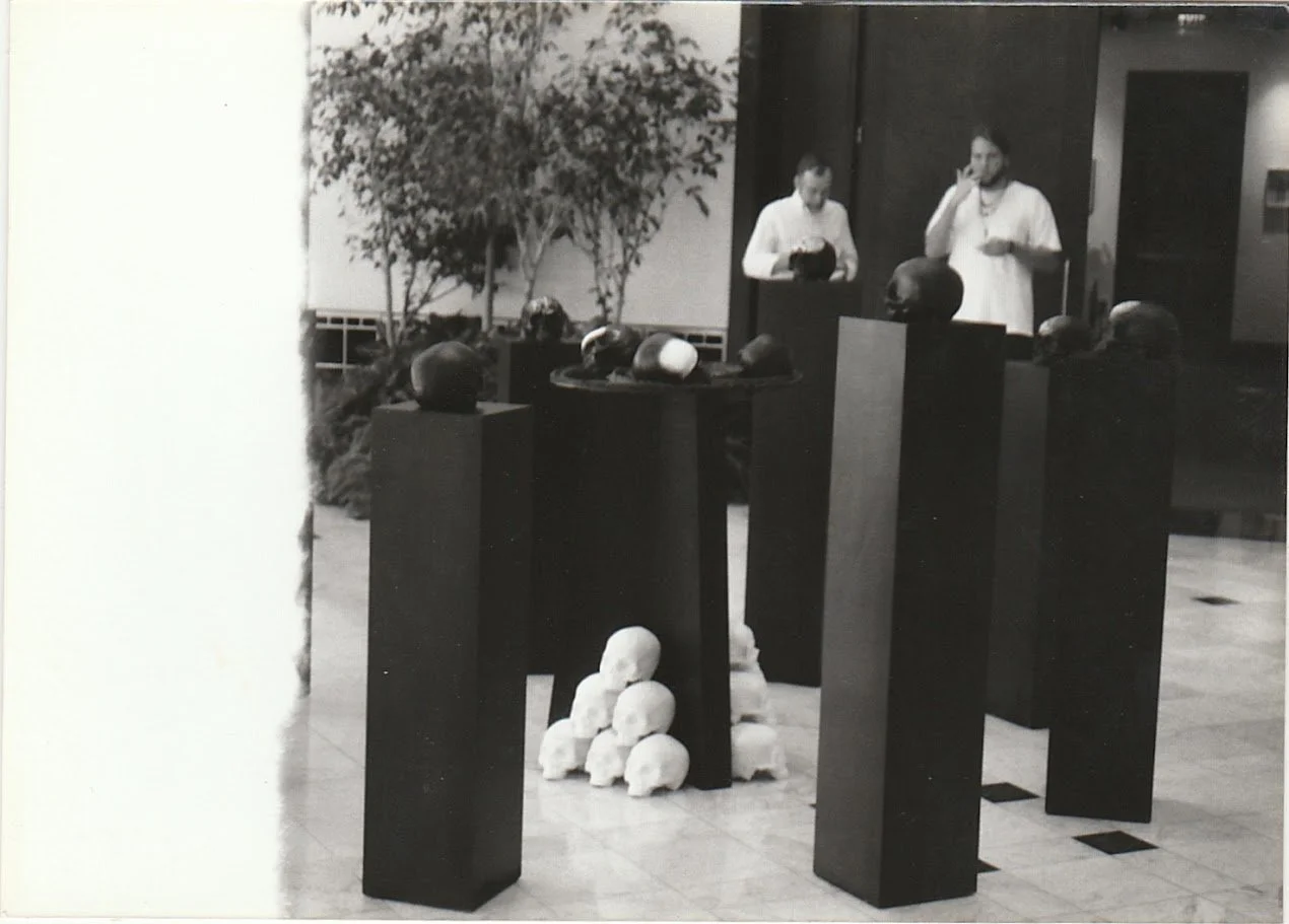 A gallery installation featuring skull sculptures displayed on tall pedestals, with a circular arrangement of skull forms suspended at the center of the composition.