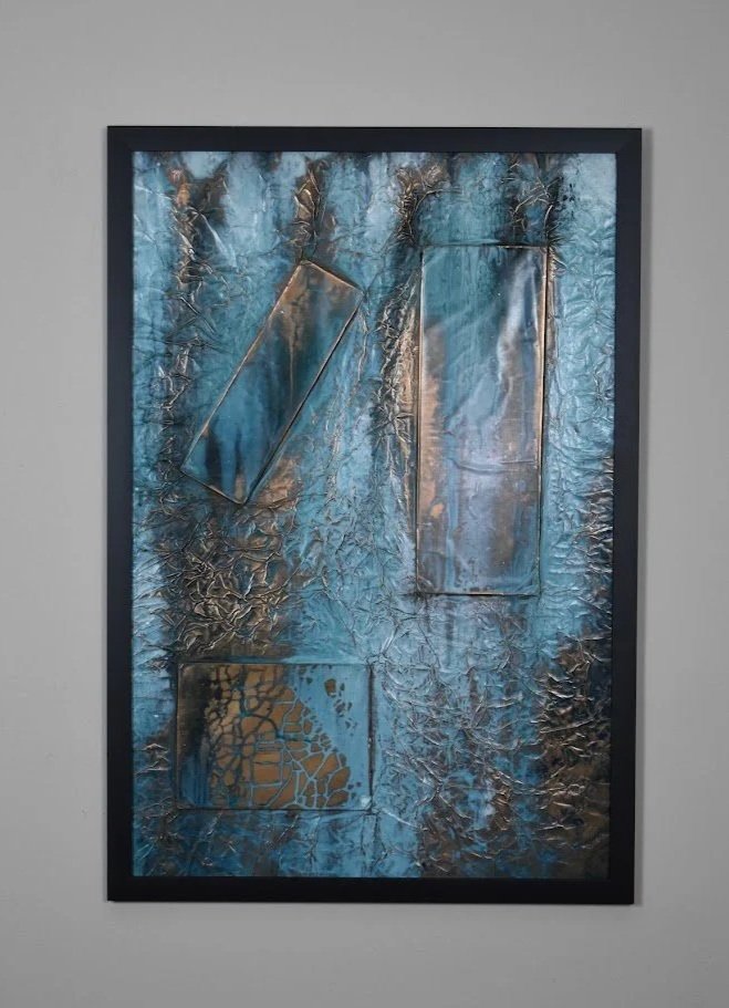Geometric forms float within a textured field of luminous blue and metallic bronze. Displayed framed on a white wall.