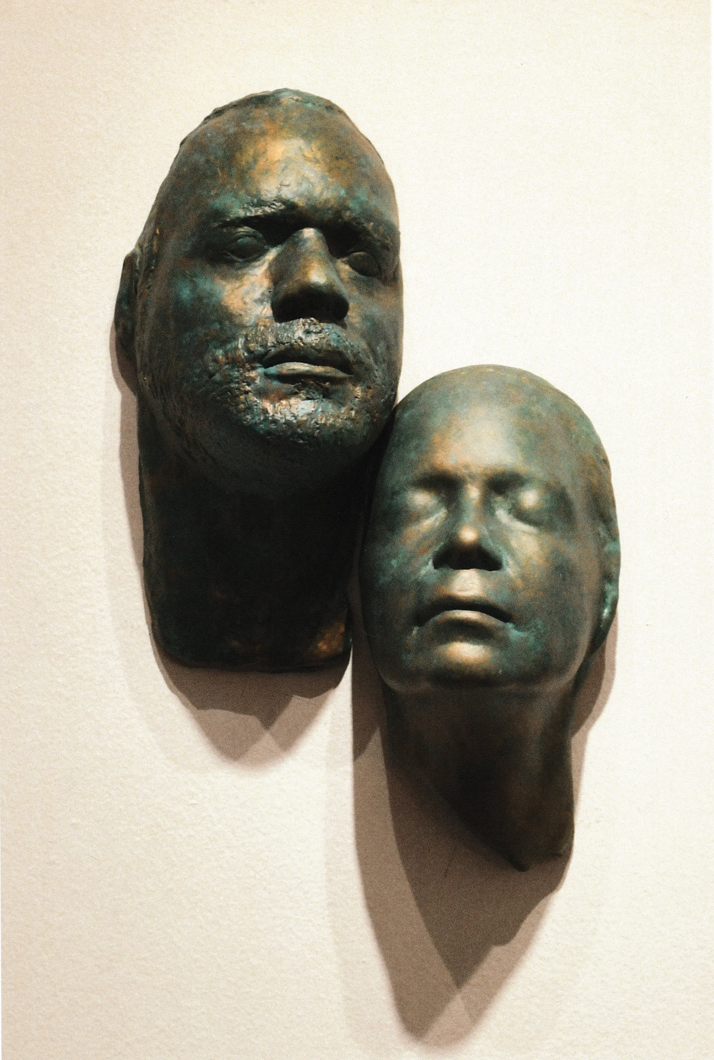 Pair of bronze life masks mounted together, one facing outward and one with eyes peacefully closed.