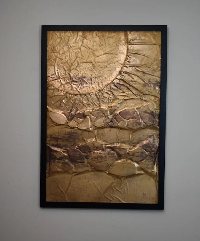 A golden orb rises above layered, fractured metallic forms. Displayed framed on a white wall.