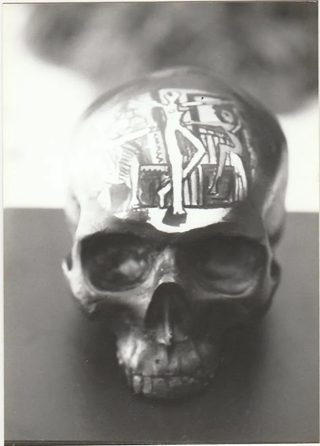 A black and white close-up view of a skull sculpture resting on a pedestal, shown from the front with deeply recessed eye sockets and a smooth, rounded cranium. The forehead contains a painting.