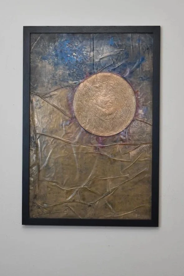 A golden orb floats within a muted field of blue, gray, and bronze. Displayed framed on a white wall.