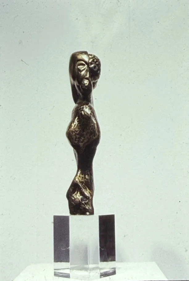 Front-facing bronze figure with simplified anatomy and exaggerated facial features, standing upright on a transparent pedestal.