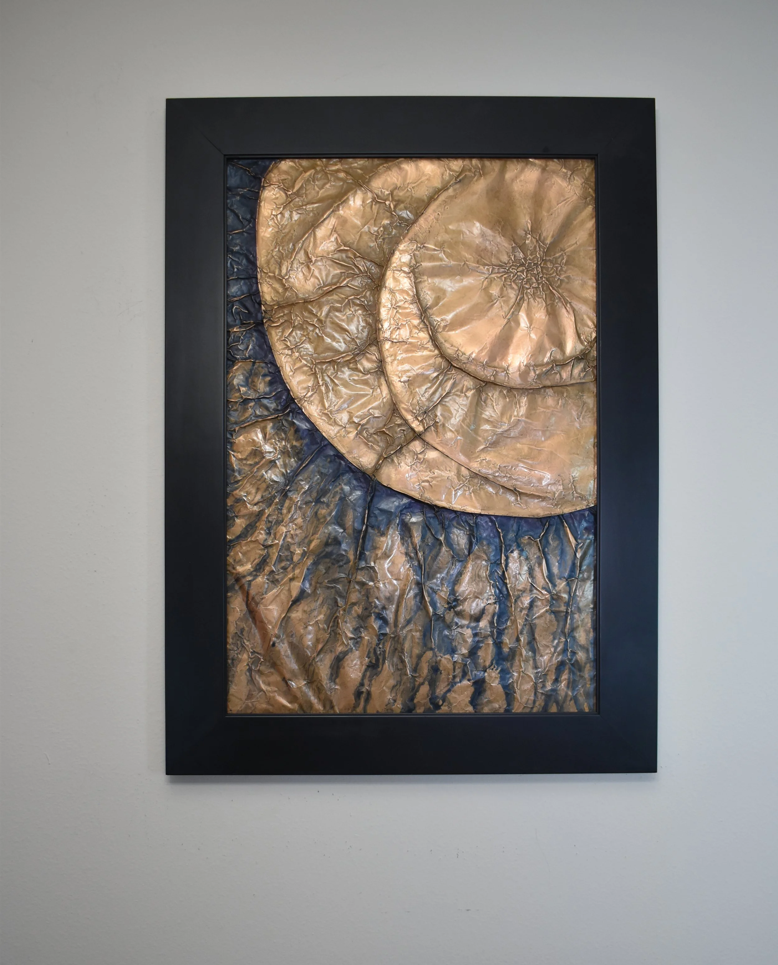 Overlapping golden orbs emerge from a deep blue metallic field. Displayed framed on a white wall.