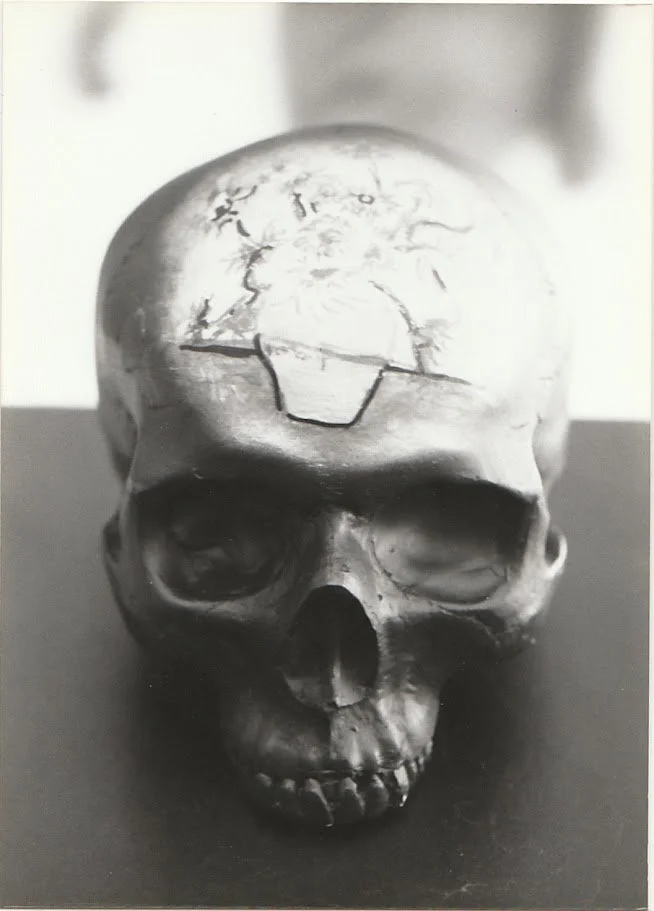 A black and white close-up view of a skull sculpture resting on a pedestal, shown from the front with deeply recessed eye sockets and a smooth, rounded cranium. The forehead contains a re-created the "Sunflowers" Picasso painting.
