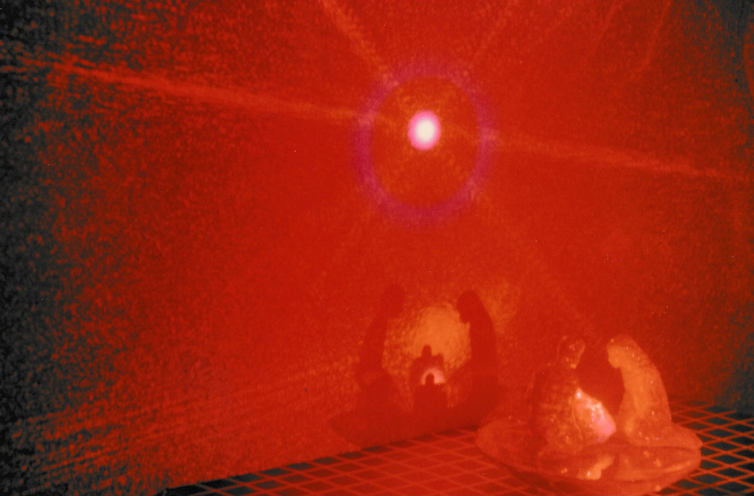 Red laser projection illuminating a sculptural arrangement on a gridded platform beneath a bright circular light source.