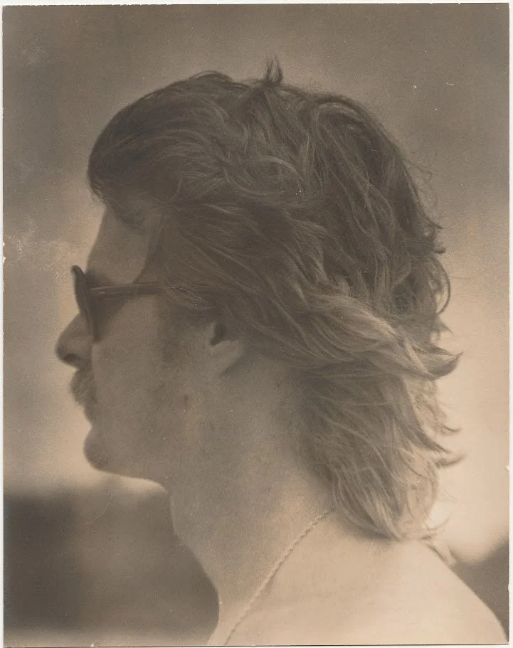 Whitney Wolf II depicted in a sepia photo from his side profile. His hair is longer, he is wearing sunglasses and has a mustache. EST 1970s