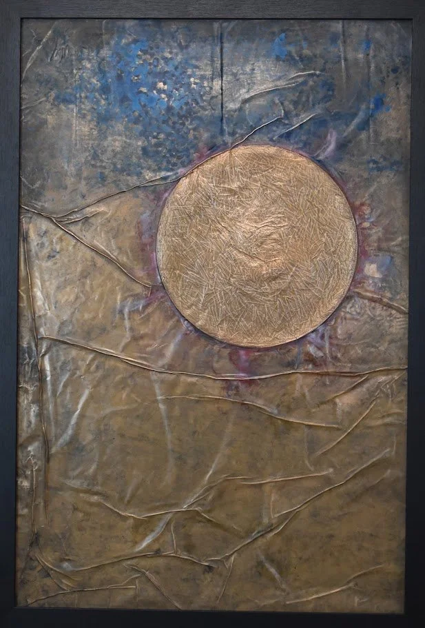 A golden orb floats within a muted field of blue, gray, and bronze. Shown up close in a black frame.