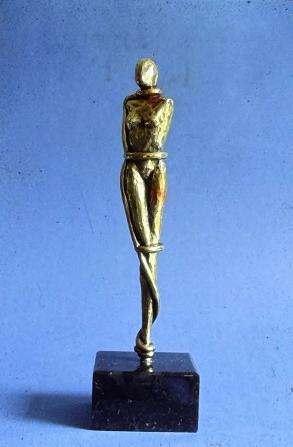 Tall stylized bronze female figure with crossed legs and folded arms. It looks as though she is abstractly bound to herself. 