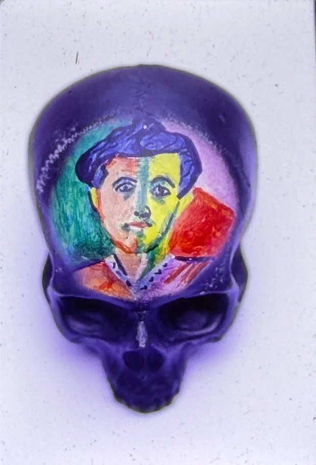 A full-color close-up view of a skull sculpture resting on a pedestal, shown from the front with deeply recessed eye sockets and a smooth, rounded cranium. The forehead contains a re-created "The Bullfighter" Picasso painting.