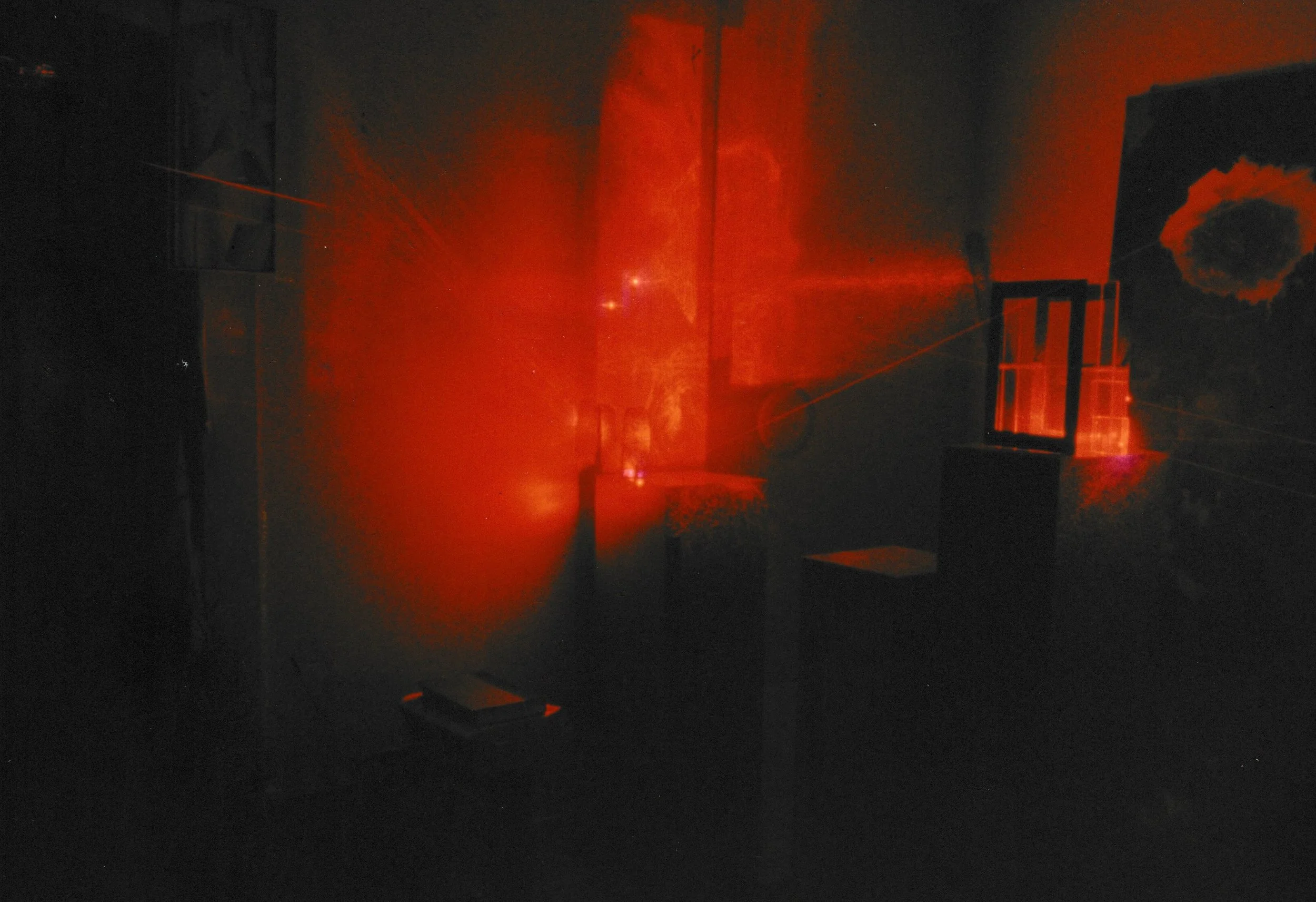 Dark gallery installation illuminated by red laser light, with glowing beams spreading across sculptural forms and pedestals.