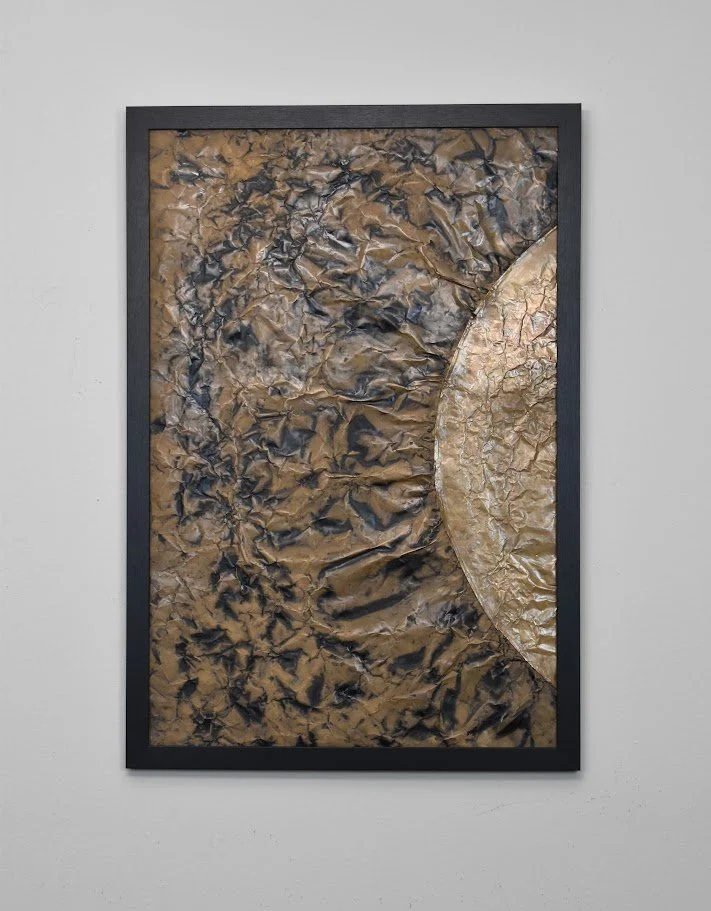 A golden orb emerges from a dark, textured metallic field. Displayed framed on a white wall.