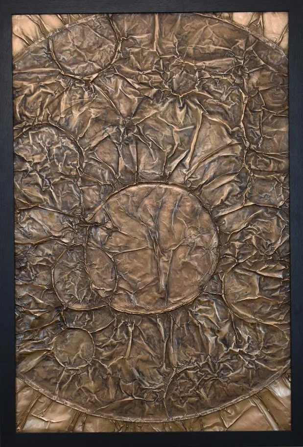 Circular forms emerge within a fractured bronze field. Shown close up in a black frame.