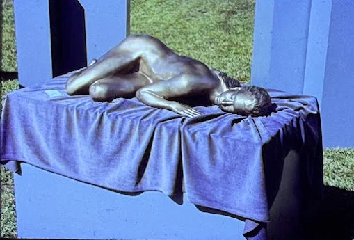 Life-size bronze reclining nude stretched across a draped platform, resting on one side with eyes closed and body relaxed.