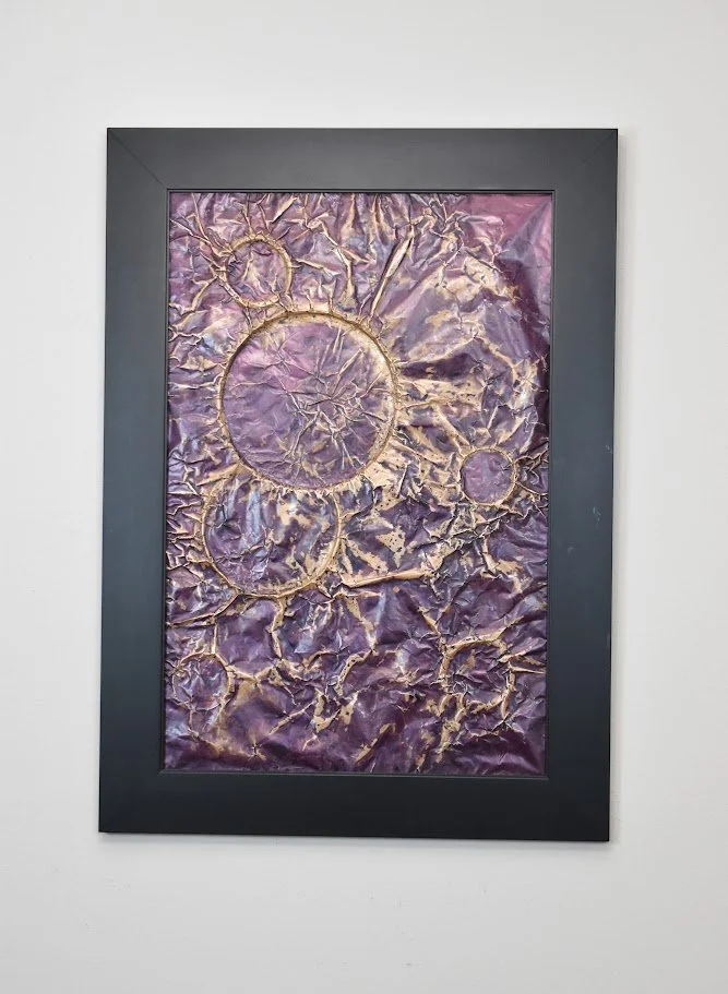 Clusters of circular forms emerge through layers of violet, lavender, and gold, resembling distant worlds suspended within a turbulent field. Displayed framed on a white wall.